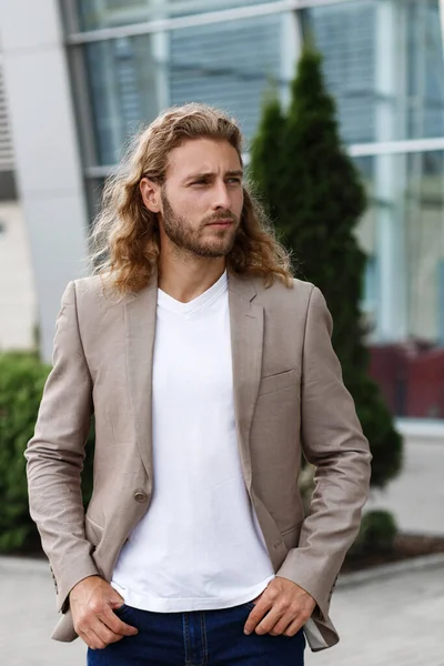 Business portrait of handsome curly smiling man with long hair in casual wear, standing on city street near business center. Confident businessman became successful