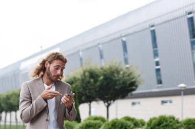 Yakışıklı, kıvırcık iş adamının portresi akıllı telefon tutarken ve gülümserken. Cep telefonu uygulamalarını, mesajları, interneti tararken, iş merkezinin yanındaki telefona bakarken başarılı bir yönetici.
