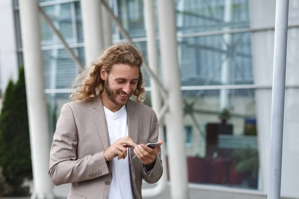 Portrait of handsome curly businessman in casual wear holding smartphone and smiling.Successful manager using mobile phone apps,texting message,browsing internet,looking at phone near business center