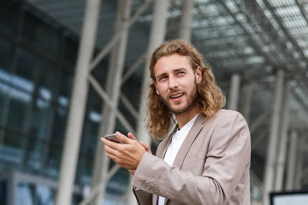 Portrait of handsome curly businessman in casual wear holding smartphone and smiling.Successful manager using mobile phone apps,texting message,browsing internet,looking at phone near business center