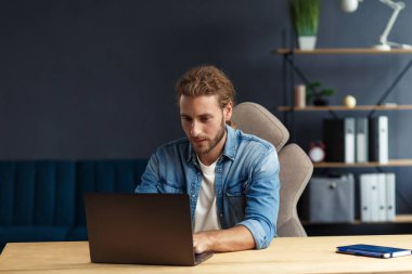 Evde dizüstü bilgisayarla çalışan uzun saçlı, kıvırcık saçlı, genç ve yakışıklı bir adam. İş yerinde oturan yakışıklı müdürün iş portresi. Çevrimiçi, çevrimiçi kurslar. İş konsepti