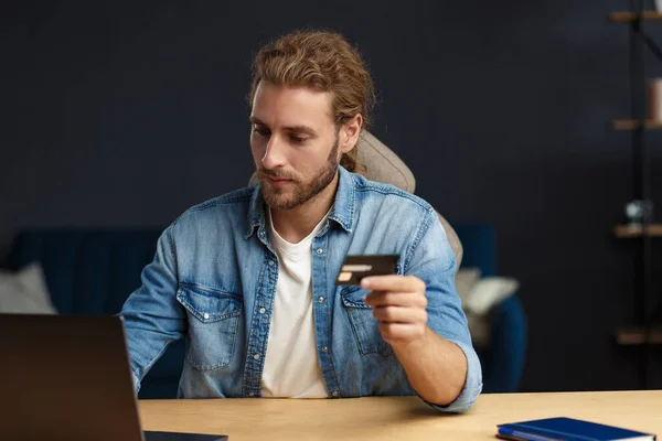 Young handsome curly smiling man with long hair in home office holding credit card and using laptop. Online shopping, online payments. Booking tickets online. Modern technologies concept