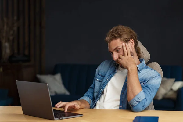 Jovem e bonito homem sorridente encaracolado com cabelo comprido trabalhando em casa escritório com laptop. Retrato de negócios de gerente bonito sentado no local de trabalho. Estudar online, cursos online. Conceito de negócio — Fotografia de Stock