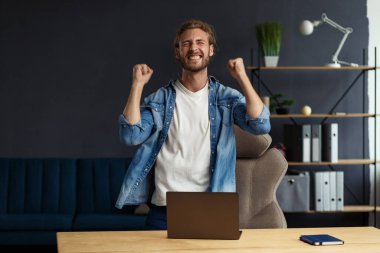 Genç yakışıklı, uzun saçlı, laptopla çalışan ve pozitifliğini ifade eden kıvırcık adam. Mutlu iş adamı olumlu haberler aldı. Ödüllendirildi, yeni sözleşme, iyi anlaşma, başarılı pazarlıklar, en iyi gün.