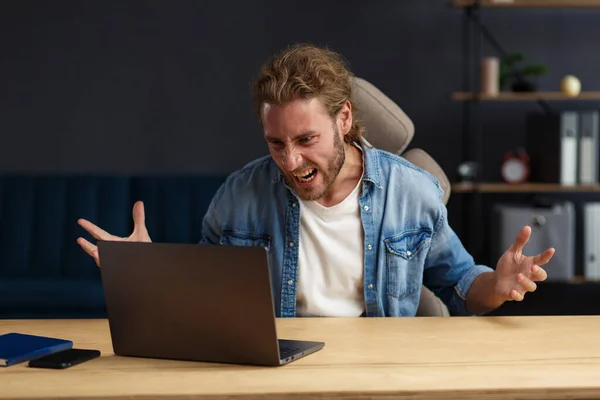 Angry young handsome curly man using laptop. Stressed freelancer lost data on broken not working laptop. Receiving bad news or email, spam message. Young people working with mobile devices