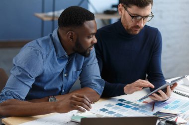 Çok ırklı mühendis ekibi tablet bilgisayarda çalışıyor. Mimarlar bina renklerini renk örnekleri, çizim ve inşaat projeleri kullanarak seçerler. İş yerinde tartışan ortaklar