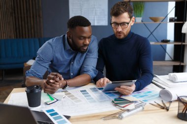 Çok ırklı mühendis ekibi tablet bilgisayarda çalışıyor. Mimarlar bina renklerini renk örnekleri, çizim ve inşaat projeleri kullanarak seçerler. İş yerinde tartışan ortaklar