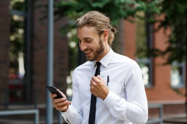 Heyecanlı ve mutlu iş adamı elinde Victoria 'dan bir akıllı telefon tutuyor. İş merkezinin arka planında beyaz gömlekli kıvırcık saçlı genç bir iş adamının portresi..