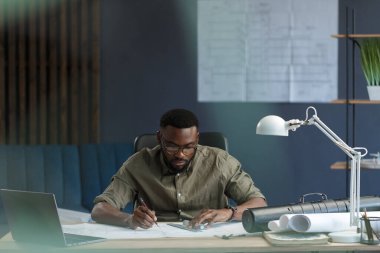 Ofiste projelerle çalışan Afro-Amerikalı bir mimar. Mühendis mimari planı inceliyor, bir inşaat projesi çiziyor. İş yerinde oturan siyah yakışıklı adamın portresi. İş konsepti