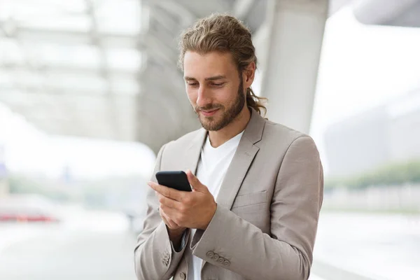Portrait of handsome curly businessman in casual wear holding smartphone and smiling.Successful manager using mobile phone apps,texting message,browsing internet,looking at phone near business center