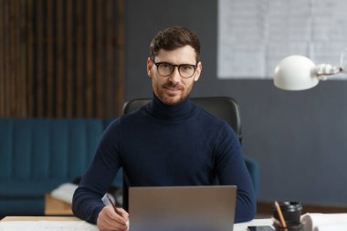 Genç iş adamı dizüstü bilgisayarla ofiste çalışıyor. İş yerinde oturan gözlüklü yakışıklı sakallı adamın iş portresi. Kendinden emin iş adamı başarılı oldu. İş konsepti