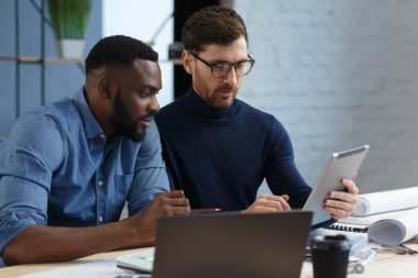 Çok ırklı mühendis ekibi tablet bilgisayarda çalışıyor. Mimarlar bina renklerini renk örnekleri, çizim ve inşaat projeleri kullanarak seçerler. İş yerinde tartışan ortaklar