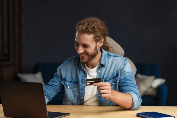 Young handsome curly smiling man with long hair in home office holding credit card and using laptop. Online shopping, online payments. Booking tickets online. Modern technologies concept — Stock Photo, Image