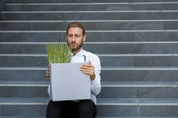 An unhappy young manager in a white shirt is fired from his job. A sad worker sits on the steps of a business center after layoffs. Crisis and unemployment