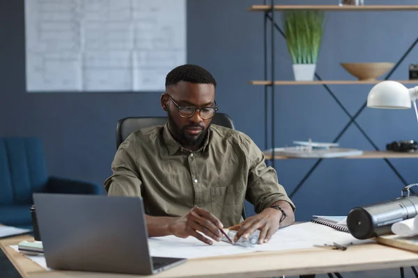 Arquitecto afro-americano que trabaja en oficina con planos. Ingeniero inspecciona plano arquitectónico, dibujando un proyecto de construcción. Retrato de un hombre guapo negro sentado en el lugar de trabajo. Concepto empresarial —  Fotos de Stock