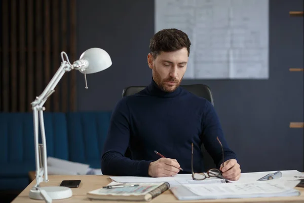 Arquitecto trabajando en la oficina con planos. Ingeniero inspeccionar plan arquitectónico, bosquejando un proyecto de construcción. Retrato de hombre barbudo guapo sentado en el lugar de trabajo. Concepto de construcción empresarial — Foto de Stock