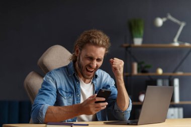 Genç yakışıklı, uzun saçlı, laptopla çalışan ve pozitifliğini ifade eden kıvırcık adam. Mutlu iş adamı olumlu haberler aldı. Ödüllendirildi, yeni sözleşme, iyi anlaşma, başarılı pazarlıklar, en iyi gün.