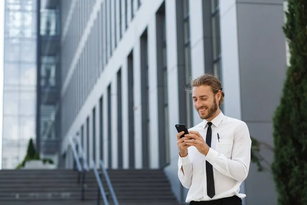 Портрет успешного молодого бизнесмена. Кудрявый мужчина в белой рубашке с телефоном на фоне современного бизнес-центра — стоковое фото