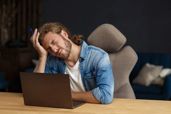 Kiégés és túlhajszolás. Fáradt üzletember túlórázik a laptoppal. Stresszes fiatal jóképű göndör férfi hosszú haj kereső ötletek leküzdésére pénzügyi válság — Stock Fotó