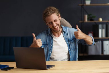 Genç yakışıklı, uzun saçlı, laptopla çalışan ve pozitifliğini ifade eden kıvırcık adam. Mutlu iş adamı olumlu haberler aldı. Ödüllendirildi, yeni sözleşme, iyi anlaşma, başarılı pazarlıklar, en iyi gün.