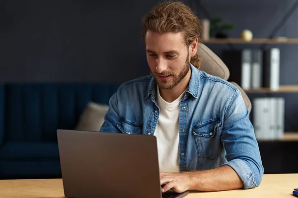 Young handsome curly smiling man with long hair working in home office with laptop. Business portrait of handsome manager sitting at workplace. Studying online, online courses. Business concept