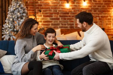 Anne babalar sevimli oğluna hediye paketi sunuyor. Noel çocukları, çocuk, portre. Mutlu bir aile Noel sabahı birlikte geçirir. Kış tatilleri, Noel kutlamaları, yeni yıl konsepti.
