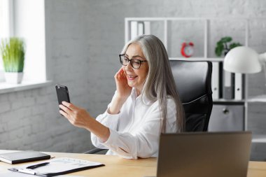 Kıdemli gri saçlı güzel kadın akıllı telefon kullanıyor ve gülümsüyor. Mutlu iş kadını cep telefonu uygulamaları kullanıyor, mesaj atıyor, internette geziniyor, internetten alışveriş yapıyor. Mobil aygıtları olan olgun insanlar