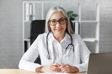 Ofisindeki yaşlı gri saçlı kadın doktorun portresi bilgisayarla bir hastayla görüntülü konuşma yapıyor. Teşhis ve tedavi tavsiyesi için doktorla çevrimiçi istişare. TeleHealth konsepti