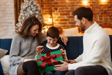 Anne babalar sevimli oğluna hediye paketi sunuyor. Noel çocukları, çocuk, portre. Mutlu bir aile Noel sabahı birlikte geçirir. Kış tatilleri, Noel kutlamaları, yeni yıl konsepti.