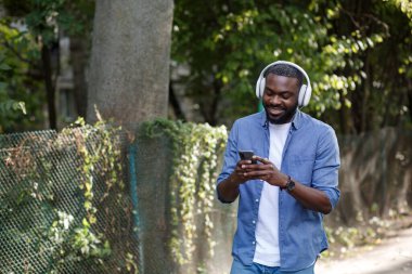 Kulaklıklı ve cep telefonlu gülümseyen adamın portresi. Kulaklıklı genç Afro-Amerikalı adam müzik uygulaması kullanarak akıllı telefondan müzik dinliyor. Sokakta yürümek, rahatlamak, rahatlamak.