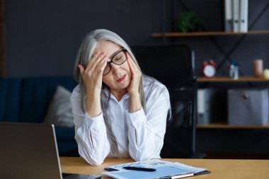 Bitkin ve aşırı çalışma kavramı. Ofiste fazla mesai yapan yorgun iş kadını. Kafası ağrıyan gri saçlı yaşlı bir kadının portresi. Dizüstü bilgisayarla çalıştıktan sonra göz yorgunluğu.