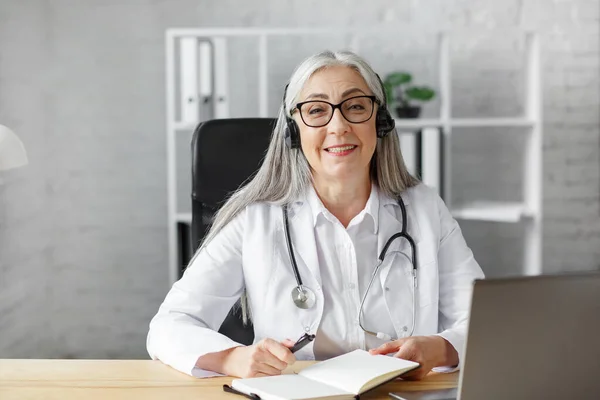 Portrait d'une femme médecin chevelue aux cheveux gris dans son bureau utilisant un ordinateur portable pour le chat vidéo avec une patiente. Consultation en ligne avec le médecin pour les diagnostics et les recommandations de traitement. Concept de télésanté — Photo