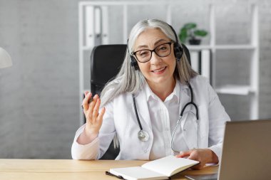 Ofisindeki yaşlı gri saçlı kadın doktorun portresi bilgisayarla bir hastayla görüntülü konuşma yapıyor. Teşhis ve tedavi tavsiyesi için doktorla çevrimiçi istişare. TeleHealth konsepti