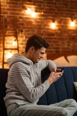 Genç serbest çalışan akıllı telefon kullanıyor ve gülümsüyor. Mutlu adam cep telefonu uygulamaları kullanıyor, mesaj yazıyor, internette geziniyor, akıllı telefona bakıyor, evde oturuyor. Mobil aygıtlarla çalışan gençler