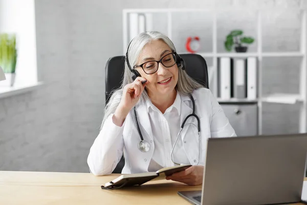 Portrait d'une femme médecin chevelue aux cheveux gris dans son bureau utilisant un ordinateur portable pour le chat vidéo avec une patiente. Consultation en ligne avec le médecin pour les diagnostics et les recommandations de traitement. Concept de télésanté — Photo