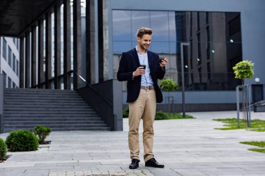 Akıllı telefon kullanan ve gülümseyen yakışıklı bir iş adamı. Mutlu genç adam cep telefonu uygulamaları kullanıyor, mesaj atıyor, internette geziniyor, akıllı telefona bakıyor. Mobil aygıtlarla çalışan gençler