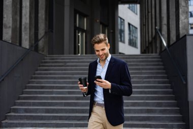 Akıllı telefonu tutan ve gülümseyen yakışıklı bir ofis çalışanı. Mutlu genç adam cep telefonu uygulamaları kullanıyor, mesaj atıyor, internette geziniyor, akıllı telefona bakıyor. İş merkezinin yakınında kahve molası.