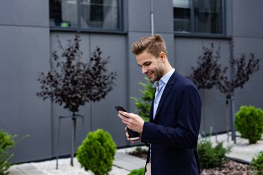 Akıllı telefonu tutan ve gülümseyen yakışıklı bir ofis çalışanı. Mutlu genç adam cep telefonu uygulamaları kullanıyor, mesaj atıyor, internette geziniyor, akıllı telefona bakıyor. İş merkezinin yakınında kahve molası.