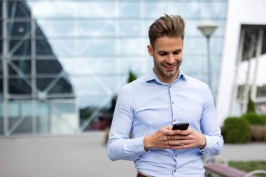 Akıllı telefon kullanan ve gülümseyen yakışıklı bir iş adamı. Mutlu genç adam cep telefonu uygulamaları kullanıyor, mesaj atıyor, internette geziniyor, akıllı telefona bakıyor. Mobil aygıtlarla çalışan gençler