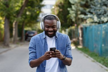 Kulaklıklı genç Afro-Amerikalı adam müzik uygulaması kullanarak akıllı telefondan müzik dinliyor. Kulaklıklı ve cep telefonlu gülümseyen adamın portresi. Sokakta yürümek, rahatlamak, rahatlamak.