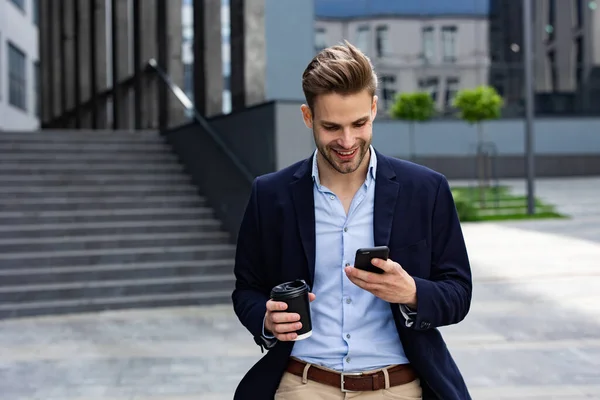 Красивый офисный работник держит смартфон и улыбается. Счастливый молодой человек использует приложения для мобильных телефонов, пишет сообщения, просматривает интернет, смотрит на смартфон. Кофе-брейк возле бизнес-центра — стоковое фото