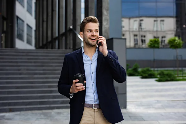Портрет красивого улыбающегося мужчины в повседневной одежде, держащего смартфон. Успешный менеджер делает звонок. Молодой бизнесмен разговаривает по телефону, стоит на городской улице рядом с бизнес-центром — стоковое фото