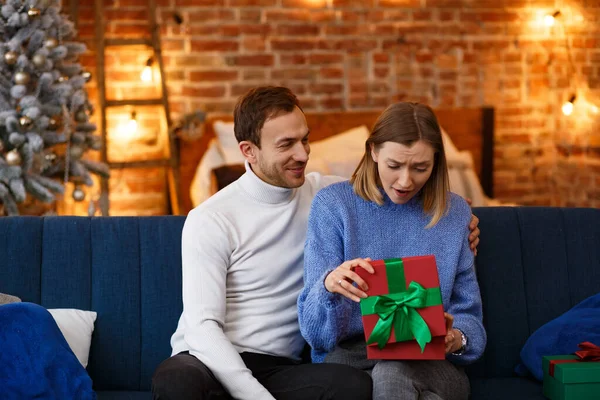 Handsome man presenting a gift to his beautiful wife and smiling. Beautiful young couple at home enjoying spending time together. Winter holidays, Christmas celebrations, New Year concept