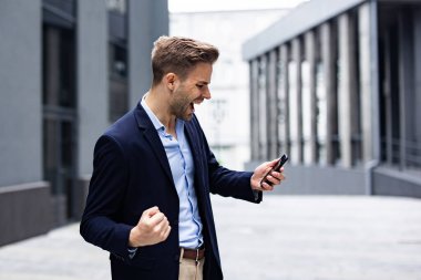 Zaferi kutlayan heyecanlı genç adam. Büyük başarı, büyük başarılar, olumlu duygular. Akıllı telefonlu yakışıklı iş adamı iyi haberler alıyor.