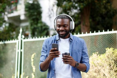 Kulaklıklı genç Afro-Amerikalı adam müzik uygulaması kullanarak akıllı telefondan müzik dinliyor. Kulaklıklı ve cep telefonlu gülümseyen adamın portresi. Sokakta yürümek, rahatlamak, rahatlamak.