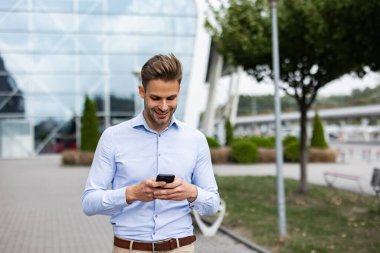 Akıllı telefonu tutan ve gülümseyen yakışıklı bir ofis çalışanı. Mutlu genç adam cep telefonu uygulamaları kullanıyor, mesaj atıyor, internette geziniyor, akıllı telefona bakıyor. İş merkezinin yakınında kahve molası.