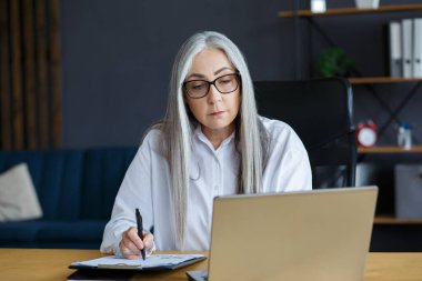 İnternette çalışan güzel, gülümseyen yaşlı bir kadının portresi. Çevrimiçi eğitim, uzaktan çalışma, evde eğitim. Not defterine yazan gri saçlı son sınıf öğrencisi. Olgun insanlar çevrimiçi kurslar öğreniyor