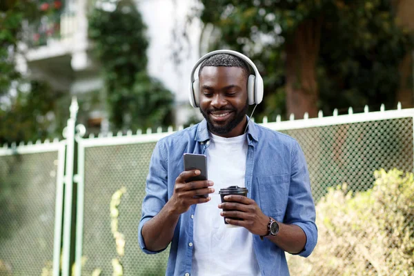 Young Afro-American man in headphones listening music on smart phone using music app. Portrait of smiling guy in earphones and mobile phone outdoors. Walking in the street, relaxation, leisure — Stock Photo, Image