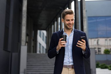 Akıllı telefonu tutan ve gülümseyen yakışıklı bir ofis çalışanı. Mutlu genç adam cep telefonu uygulamaları kullanıyor, mesaj atıyor, internette geziniyor, akıllı telefona bakıyor. İş merkezinin yakınında kahve molası.