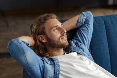 Rahatlama zamanı. Sıradan tarzda kıvırcık saçlı bir adam, evdeki kanepede oturuyor, gülümsüyor ve dinleniyor. Keyifli vakit geçirme zamanı. Evde dinlen.